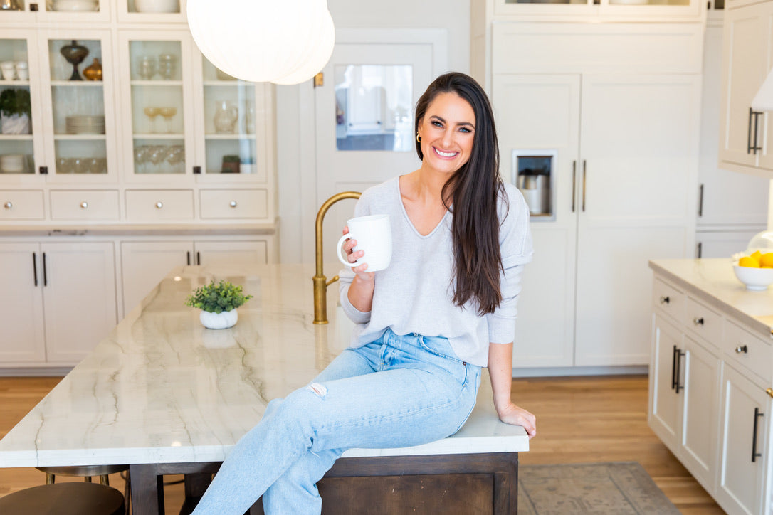 Nutrition by Gina; picture of Gina Lilley, Registered Dietitian Nutritionist sitting in a white kitchen with marble countertops holding a cup of coffee. 